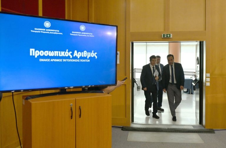 εικόνα που δείχνει σε πρώτο πλάνο μια μεγάλη οθόνη όπου αναγράφεται η φράση Προσωπικός Αριθμός από την ψηφιακή πλατφόρμα, ενώ στο φόντο διακρίνονται ο Μιχάλης Χρυσοχοΐδης αριστερά και ο Δημήτρης Παπαστεργίου δεξιά να προσέρχονται στη συνέντευξη Τύπου