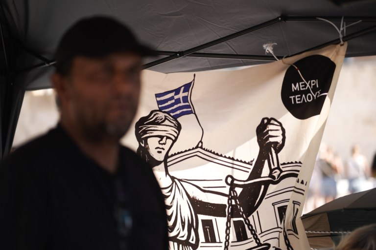 εικόνα που δείχνει σε πρώτο πλάνο τον απεργό πείνας Πάνο Ρούτσι που έχασε τον γιο του στα Τέμπη και πίσω ένα πανό με Θέμιδα και δεξιά επάνω τη φράση «μέχρι τέλους»